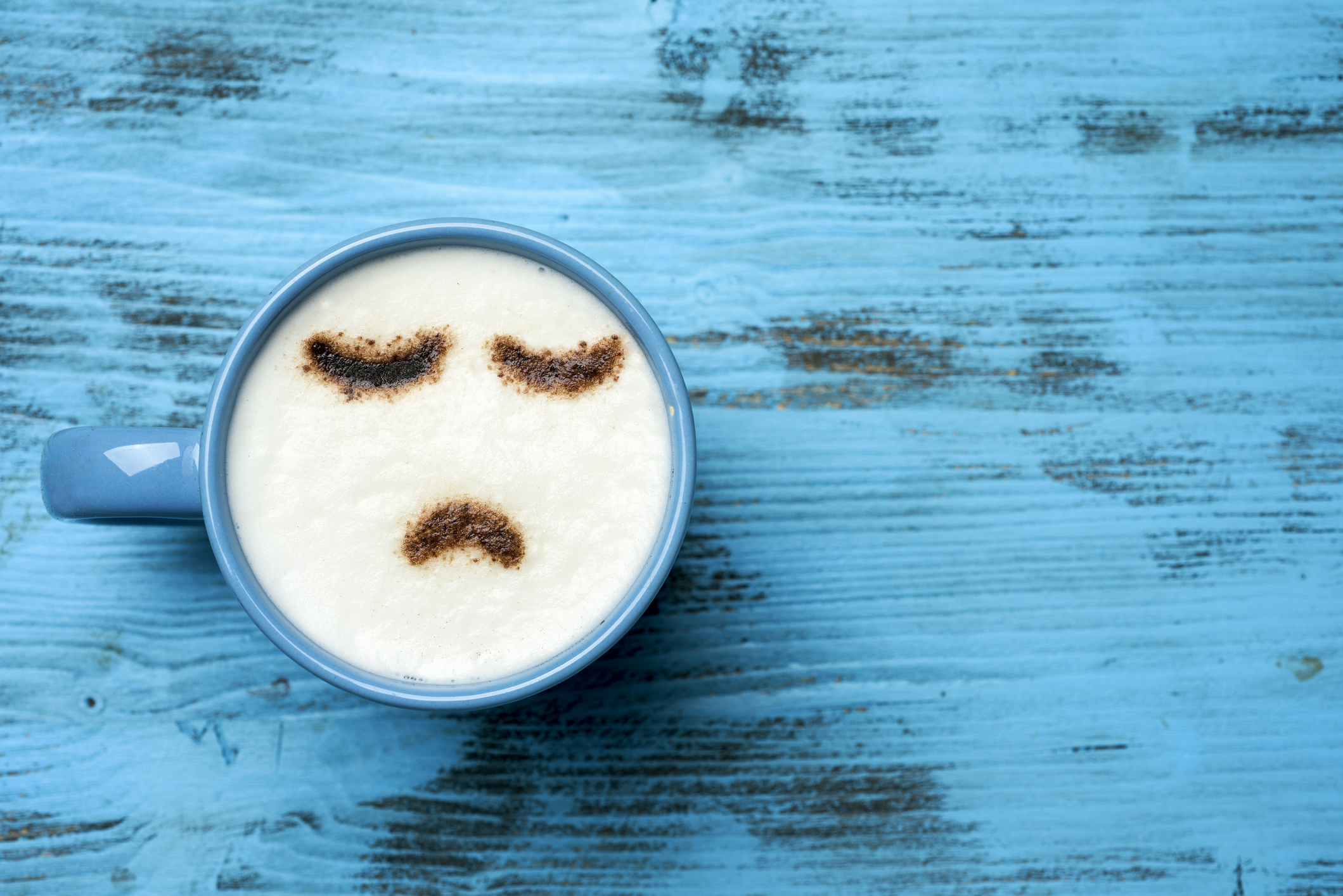 high-angle shot of a blue cup of cappuccino with a sad face drawn with cocoa powder on its milk foam, on a blue rustic table with a blank space on the right