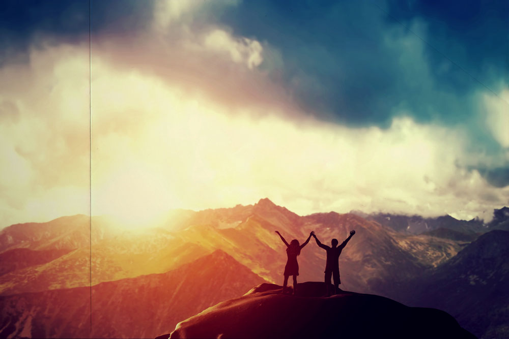 A happy couple standing together on the peak of a mountain with hands raised admiring breathtaking view at sunset