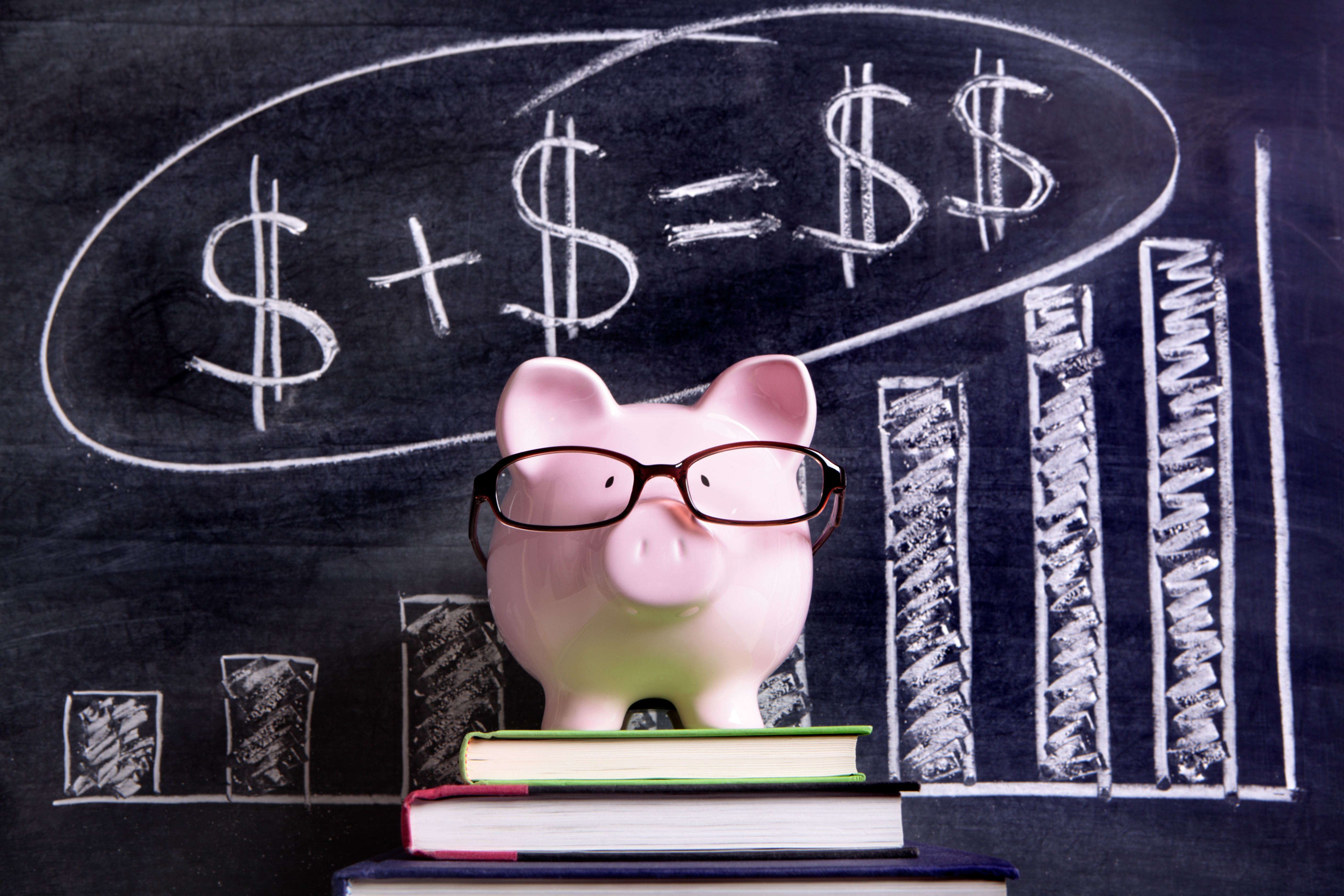 Pink piggy bank with glasses standing on books next to a blackboard with simple money math.  Sharp focus on the piggy bank with blackboard slightly blurred.