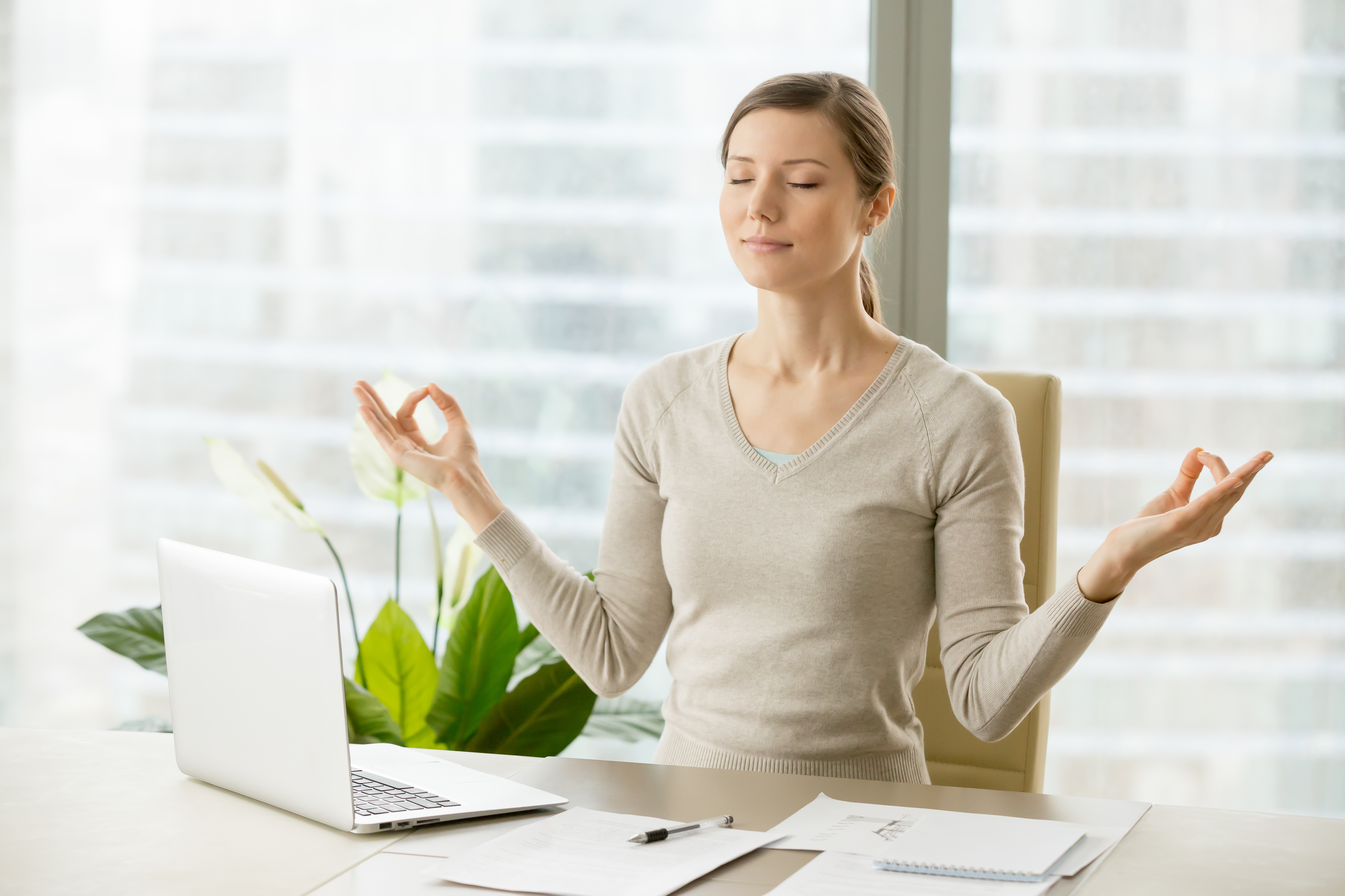 Relaxed woman meditating at workplace, practicing eastern spiritual practices for stress relief and mental health while sitting at desk in front of laptop. Short break in work for strength recovery