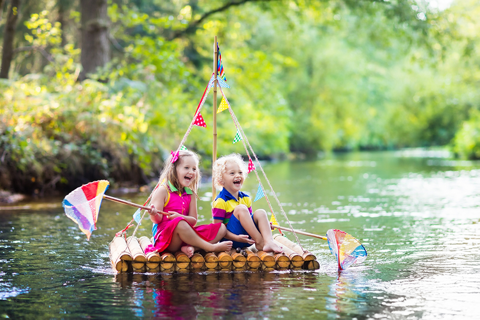 Two children on wooden raft catching fish with a colorful net in a river and playing with water on hot summer day. Outdoor fun and adventure for kids. Boy and girl in toy boat. Sailor role game.