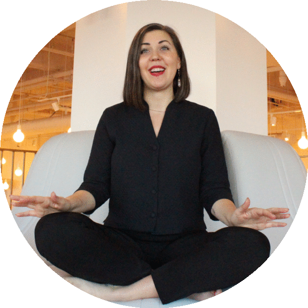 a middle aged white woman with brunette hair wearing all black clothing and red lipstick, sitting in a meditation position and smiling