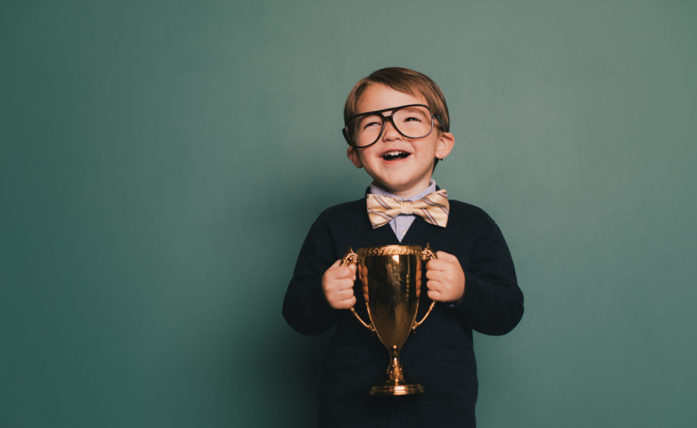 A young nerd boy wearing a bow tie and eyeglasses with a cheesy smile holds the winning contest trophy. He standing in class in front of a blackboard with his spelling bee trophy. He loves education and loves being smart.