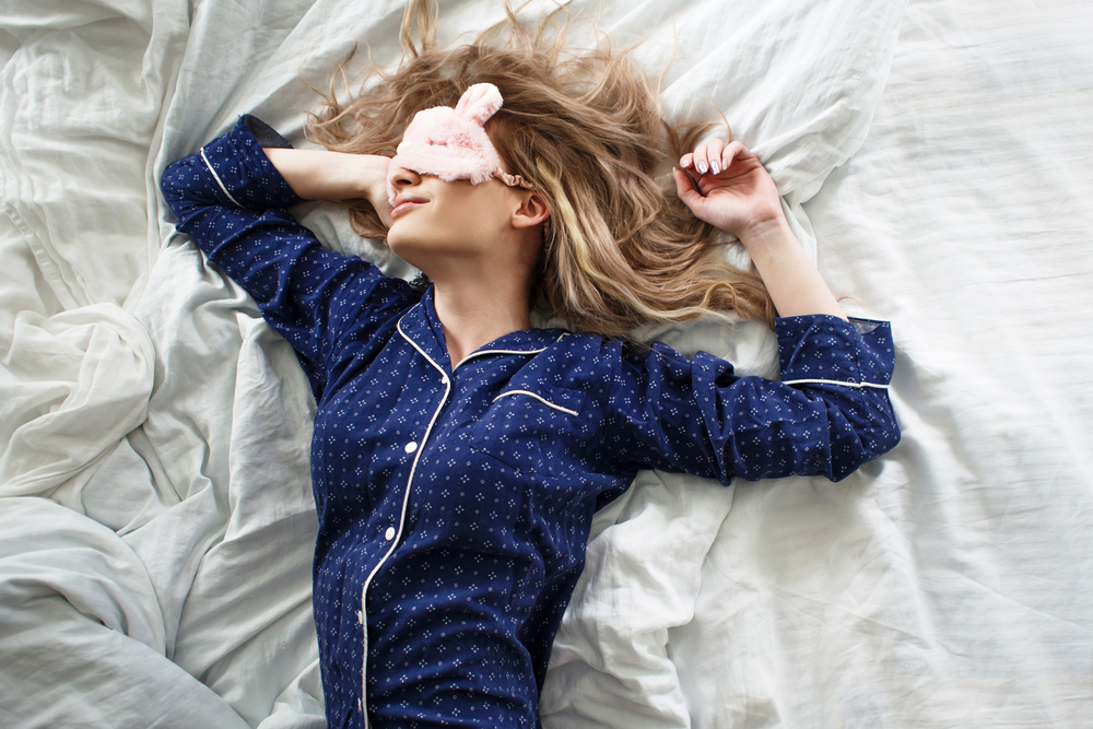 A blonde woman in blue pajamas and a pink eye mask asleep in white bed sheets.