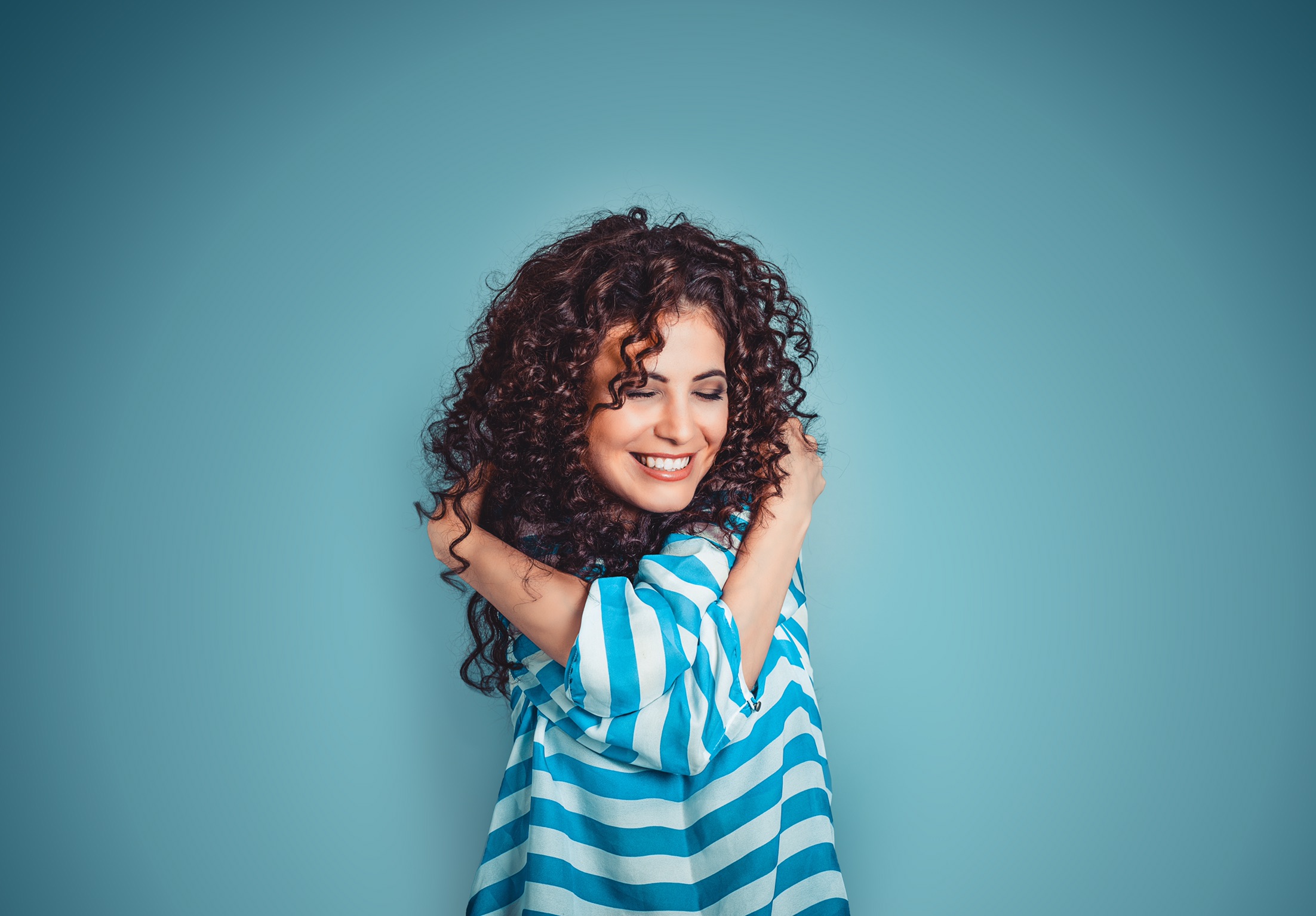 Closeup portrait confident smiling woman holding hugging herself isolated blue wall background. Positive human emotion, facial expression, feeling, reaction, situation, attitude.