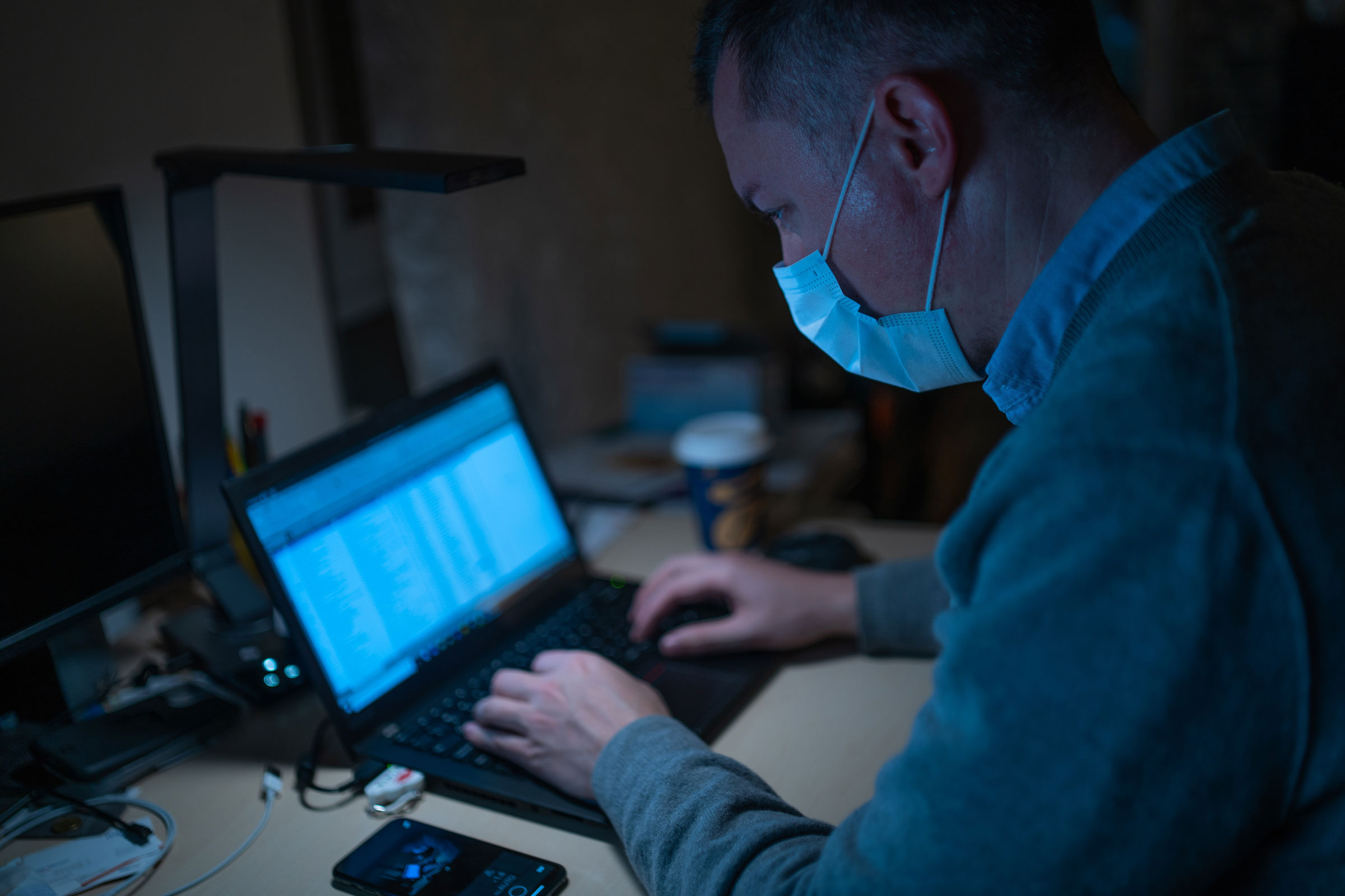 A man in a mask working on a laptop in a dimly lit room