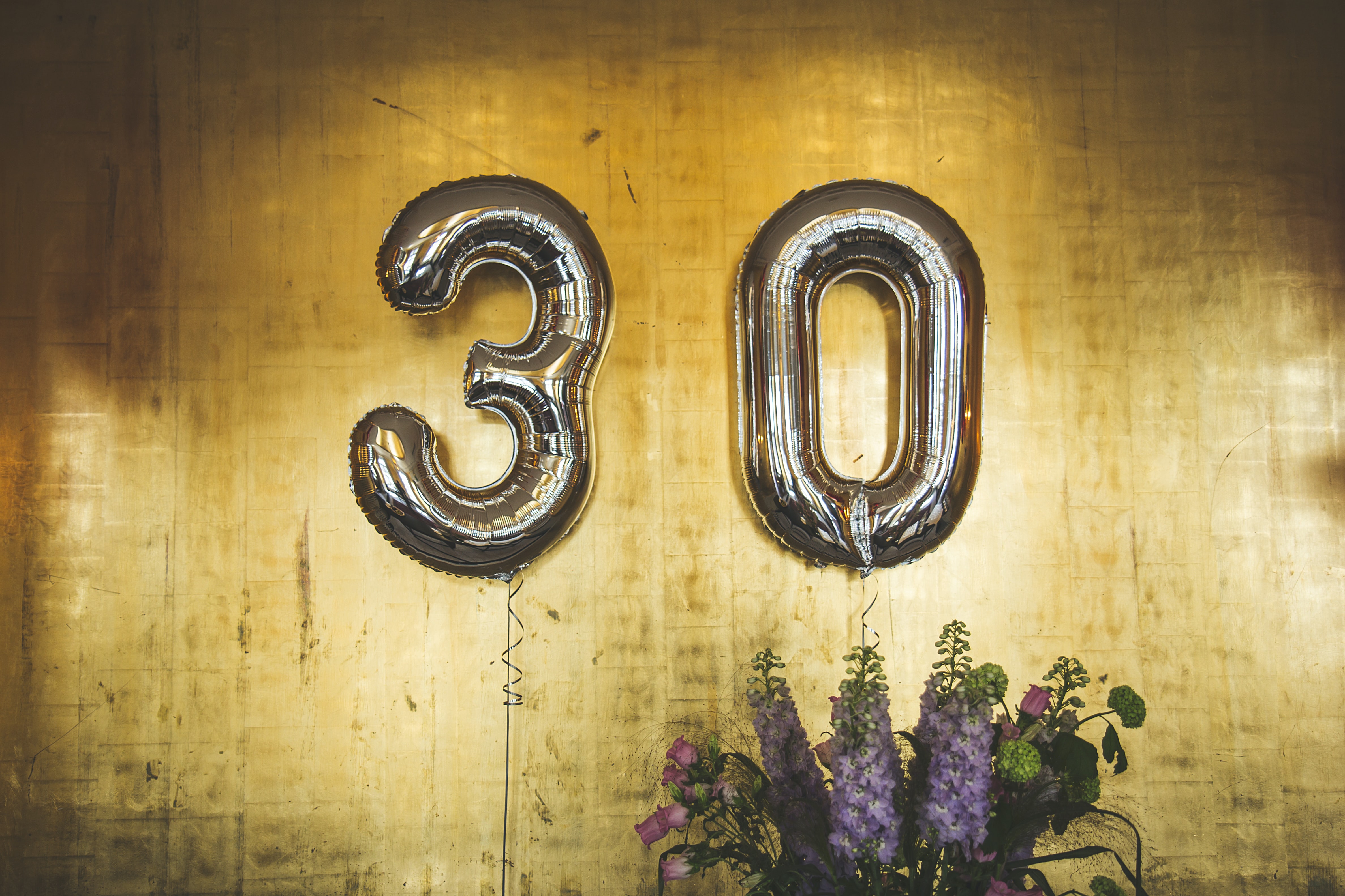 Numbered gold balloons that spell out the number 30 against a wooden backdrop and purple flowers.