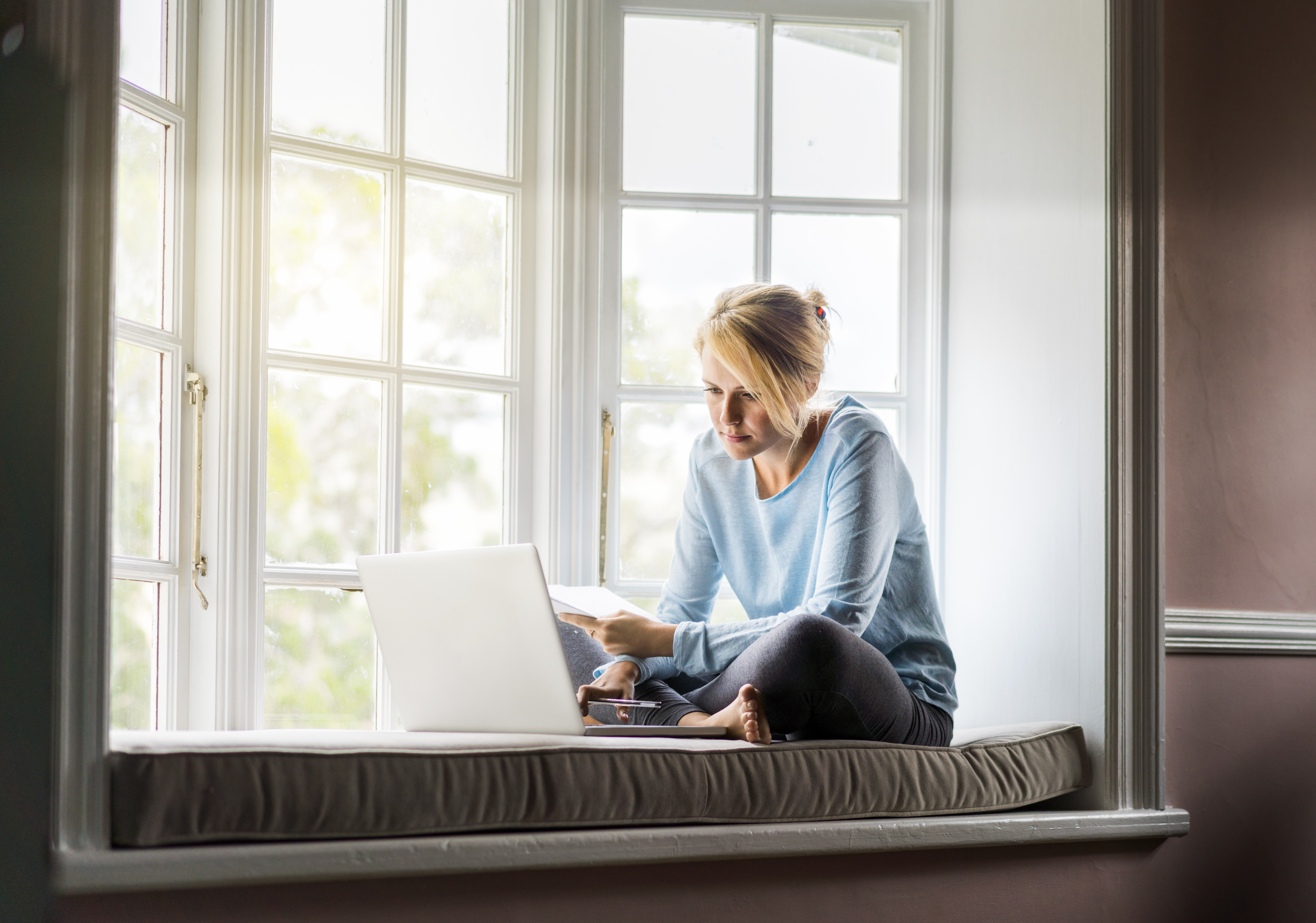 A full length photo of woman using laptop while holding documents at home. Young female is sitting on window sill. She is working from home.
