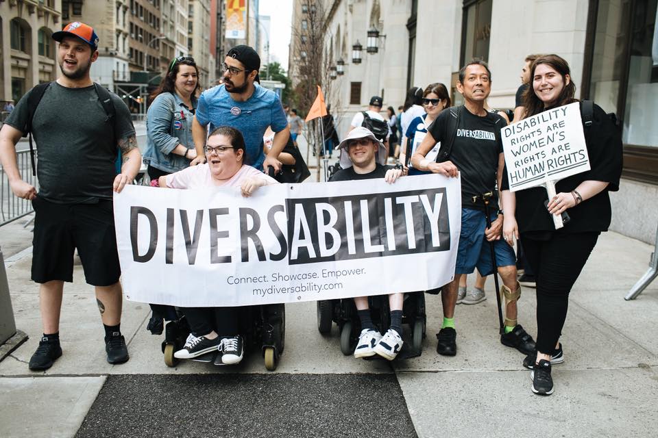 A photograph of a group of Diversability community members walking in New York City’s 2018 Disability Pride Parade. Four people are holding a Diversability banner. Another person is holding a sign that reads “disability rights are women’s rights are human rights.” This sign is written in all capital letters, and “human rights” is underlined twice.