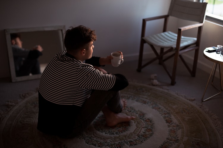 Young male sitting on a rug holding a cup of coffee looking out the window