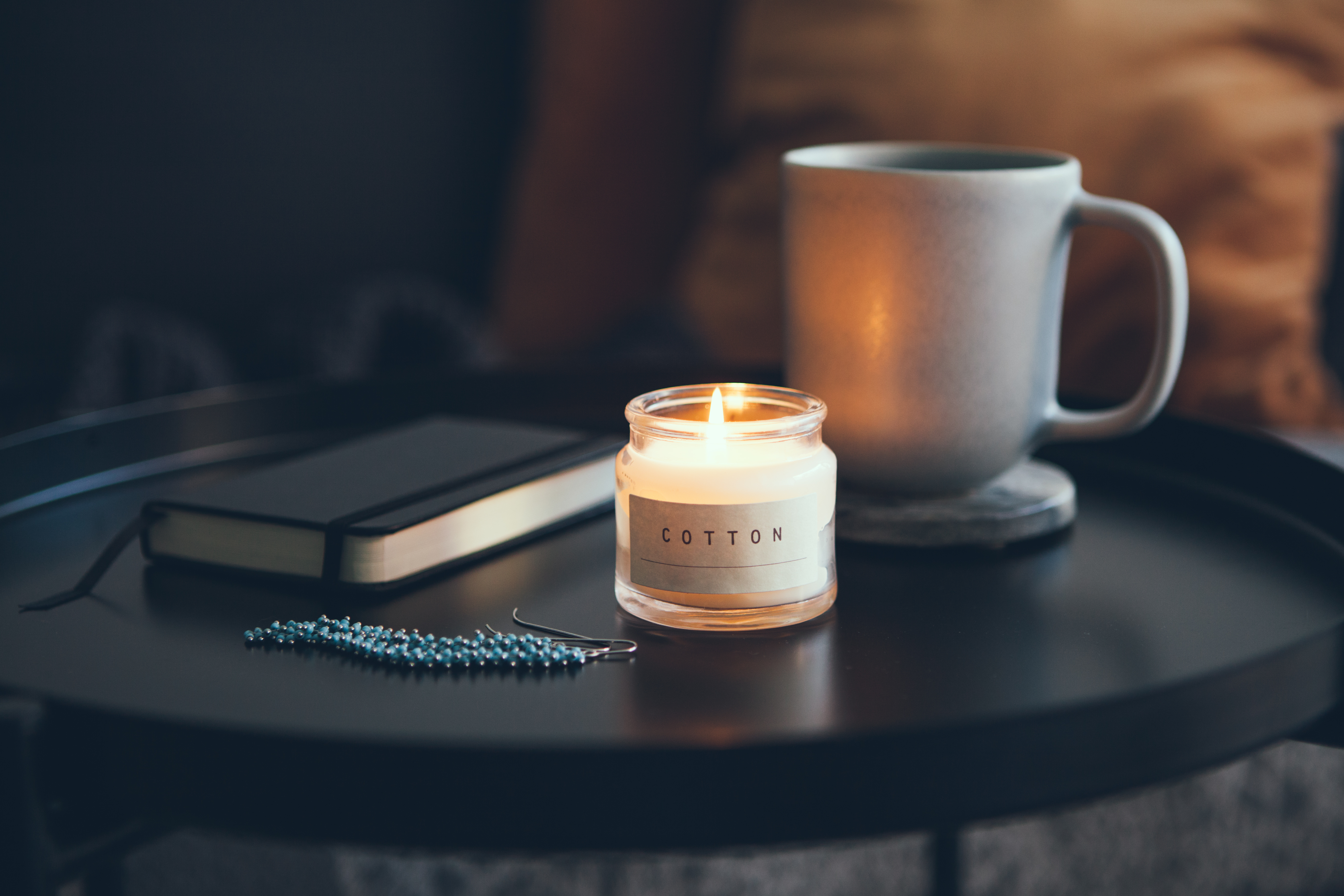 Cozy interior details. Little round table with cup of delicious tea and candle, notepad and beautiful blue color and silver earrings. Business woman resting after busy day. Selective focus.