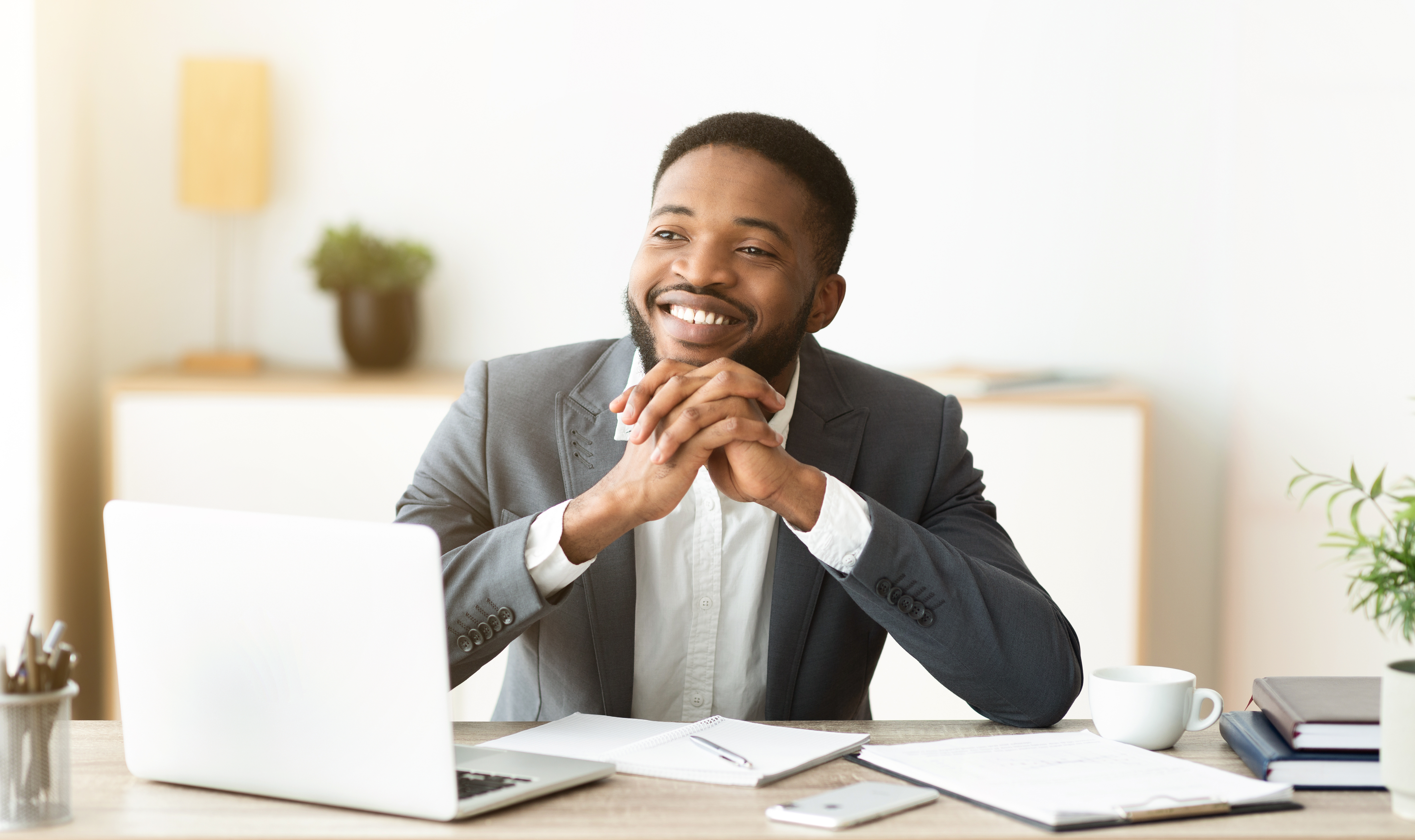 Dreamy african american businessman satisfied by results of his company, sitting at workplace in office