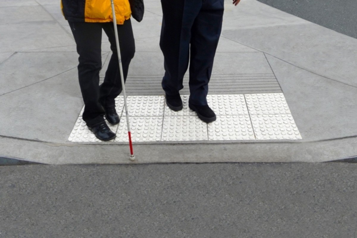 [Image Description: A picture of two people&#039;s legs. They are standing together at a crosswalk. One of these individuals is holding a white cane.] 