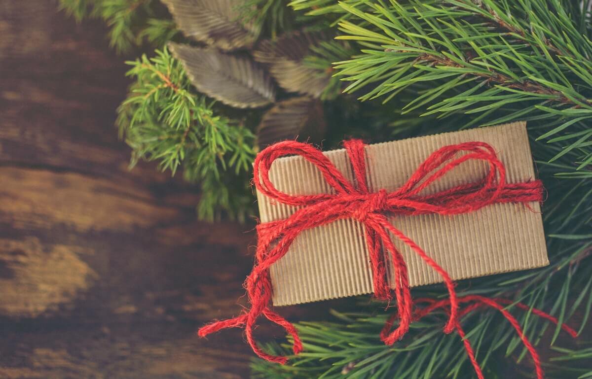 brown paper wrapped gift with red string ribbon sitting underneath Christmas tree branches