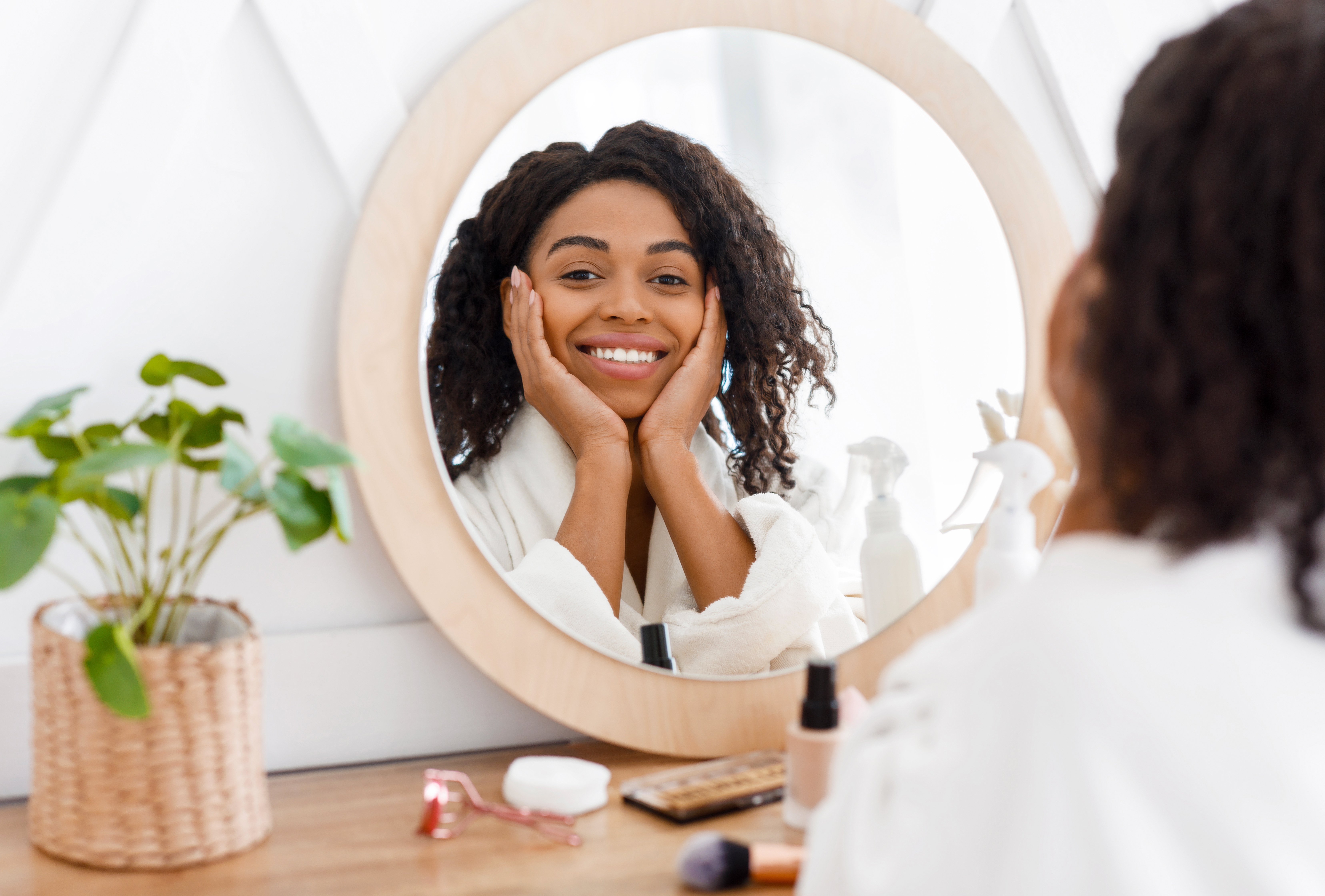 Self-Care. Beautiful African American Woman In Bathrobe Ready To Apply Makeup, Reflecting In Mirror Of Toilette Dresser Table, Looking At Camera