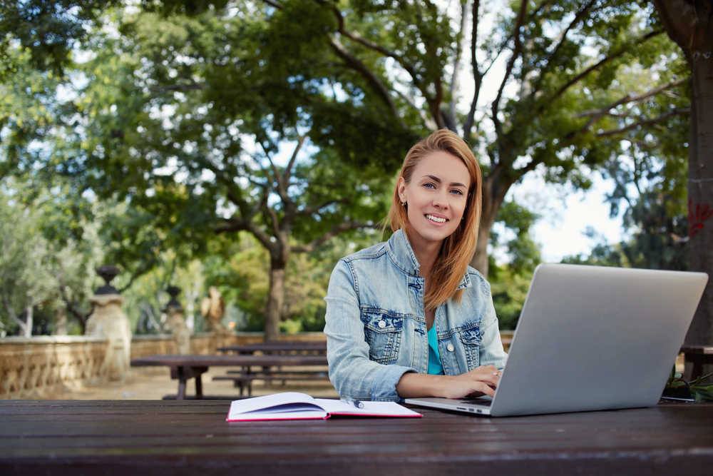 Gorgeous happy female student looking at camera while sitting with laptop computer in University park in spring day,young woman with beautiful smile posing during work on portable net-book on a campus