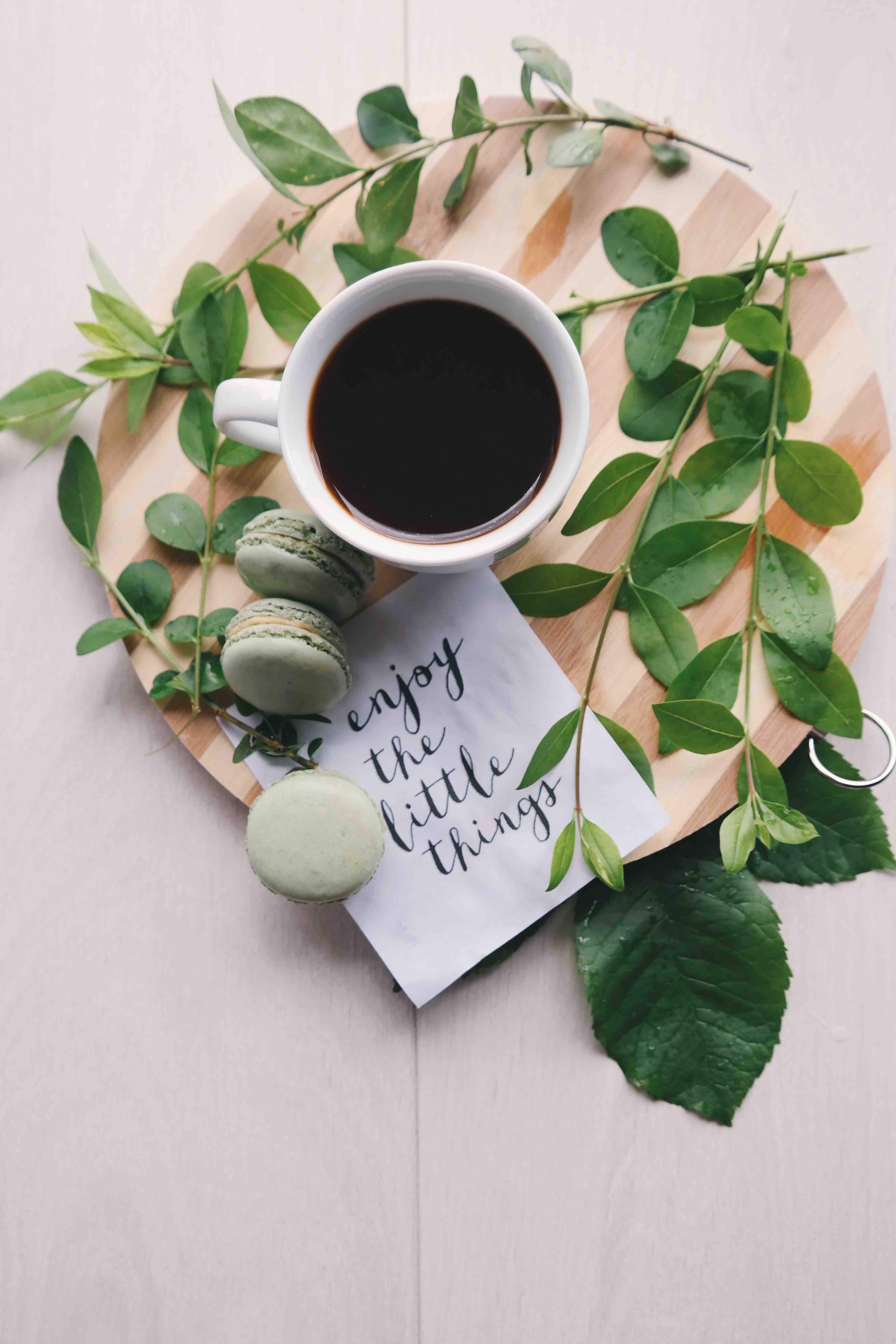 cup of coffee on a wood board, with green leaves, macarons and a napkin that says &quot;Enjoy the little things.&quot;