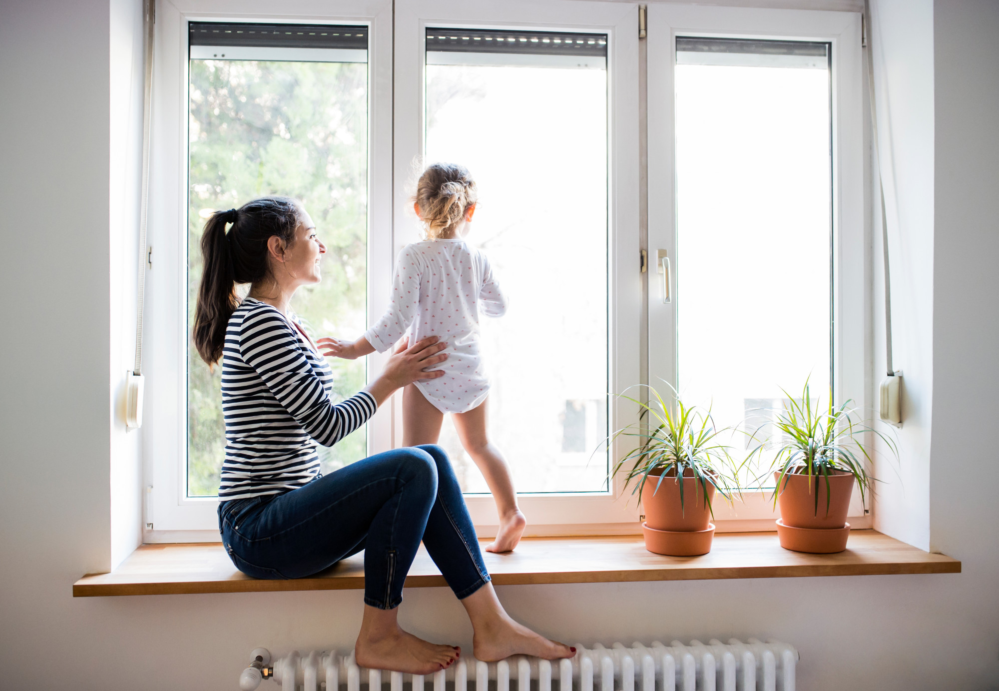 Beautiful young mother sitting on window sill with her cute little daughter looking out of window