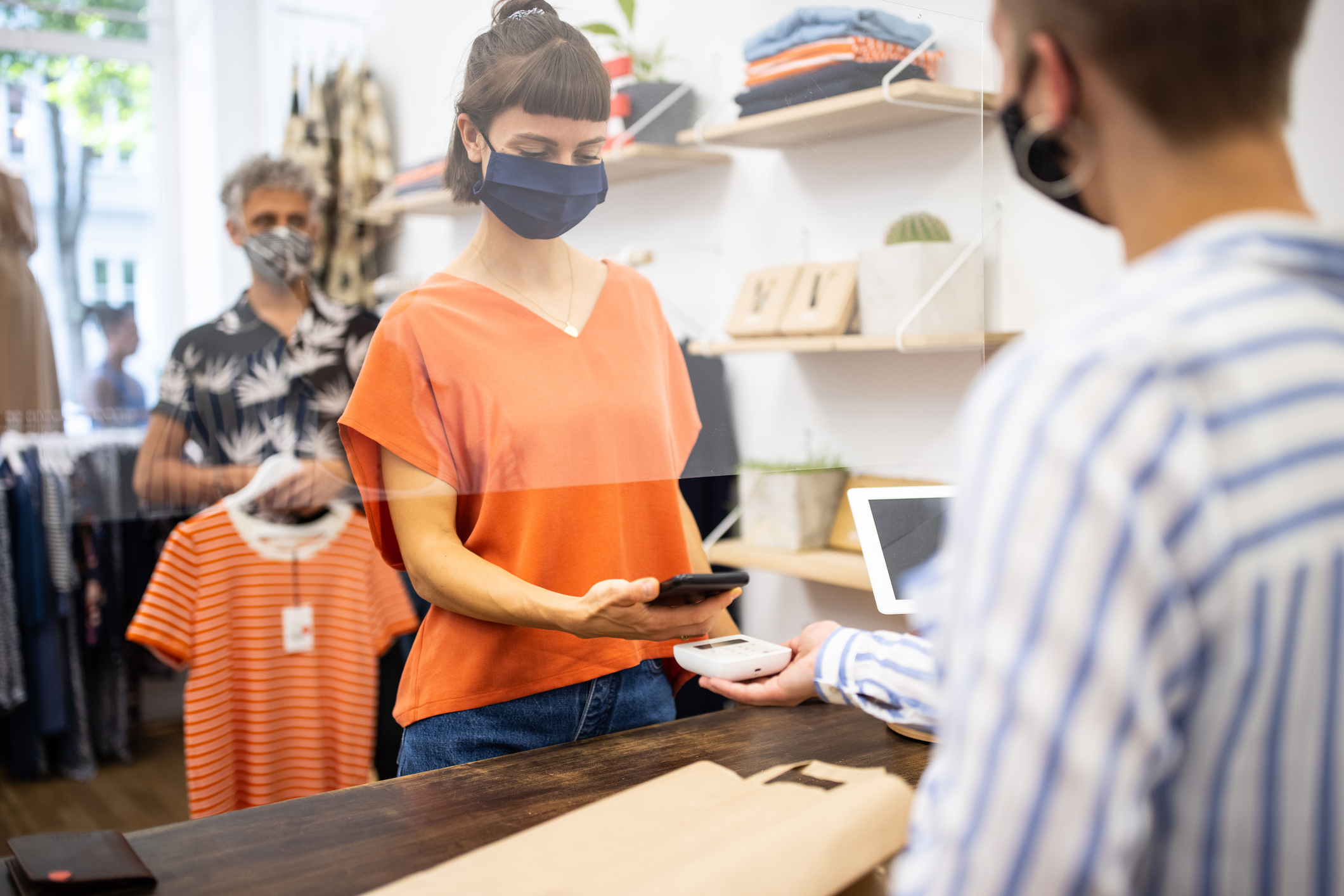 Young woman in clothing store making contactless mobile payment. Customers and store owner wearing protective face masks.