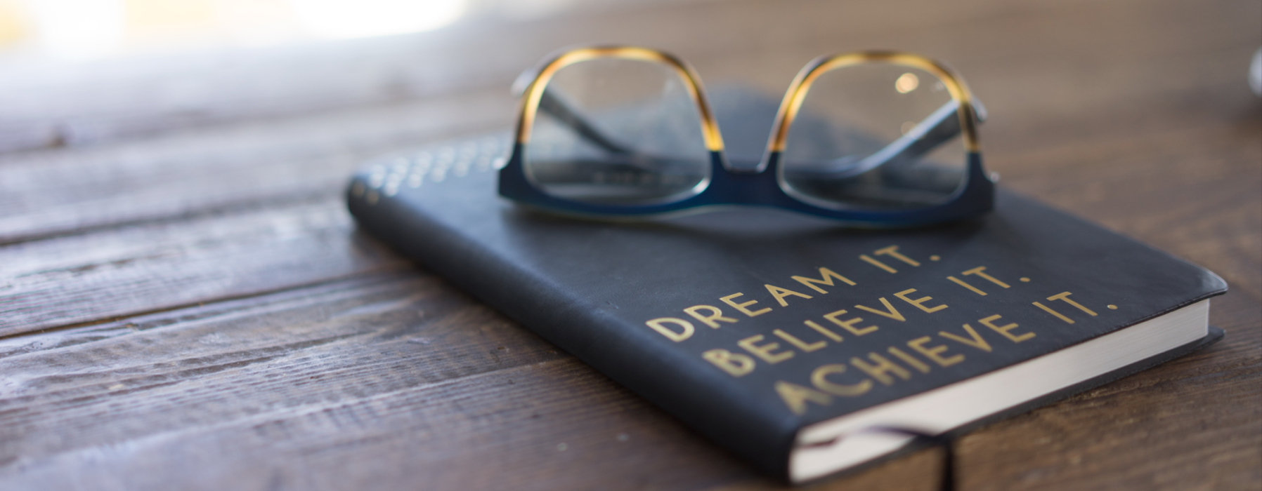 Glass on top of journal reading &quot;Dream it. Believe it. Achieve it.&quot; Photo for Unsplash by Carolyn Christine.