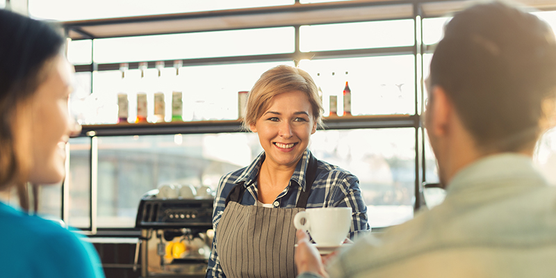 Happy smiling barista making coffee and talking to her guests at bar counter. Experienced female bartender welcoming couple. Small business, occupation people and service concept, copy space