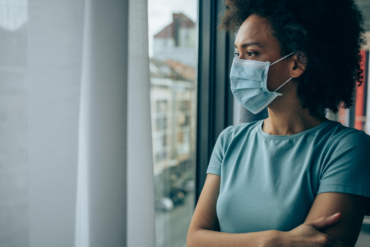 Beautiful african-american woman wearing protective face mask and looking through window at home during Coronavirus/COVID-19 pandemic.