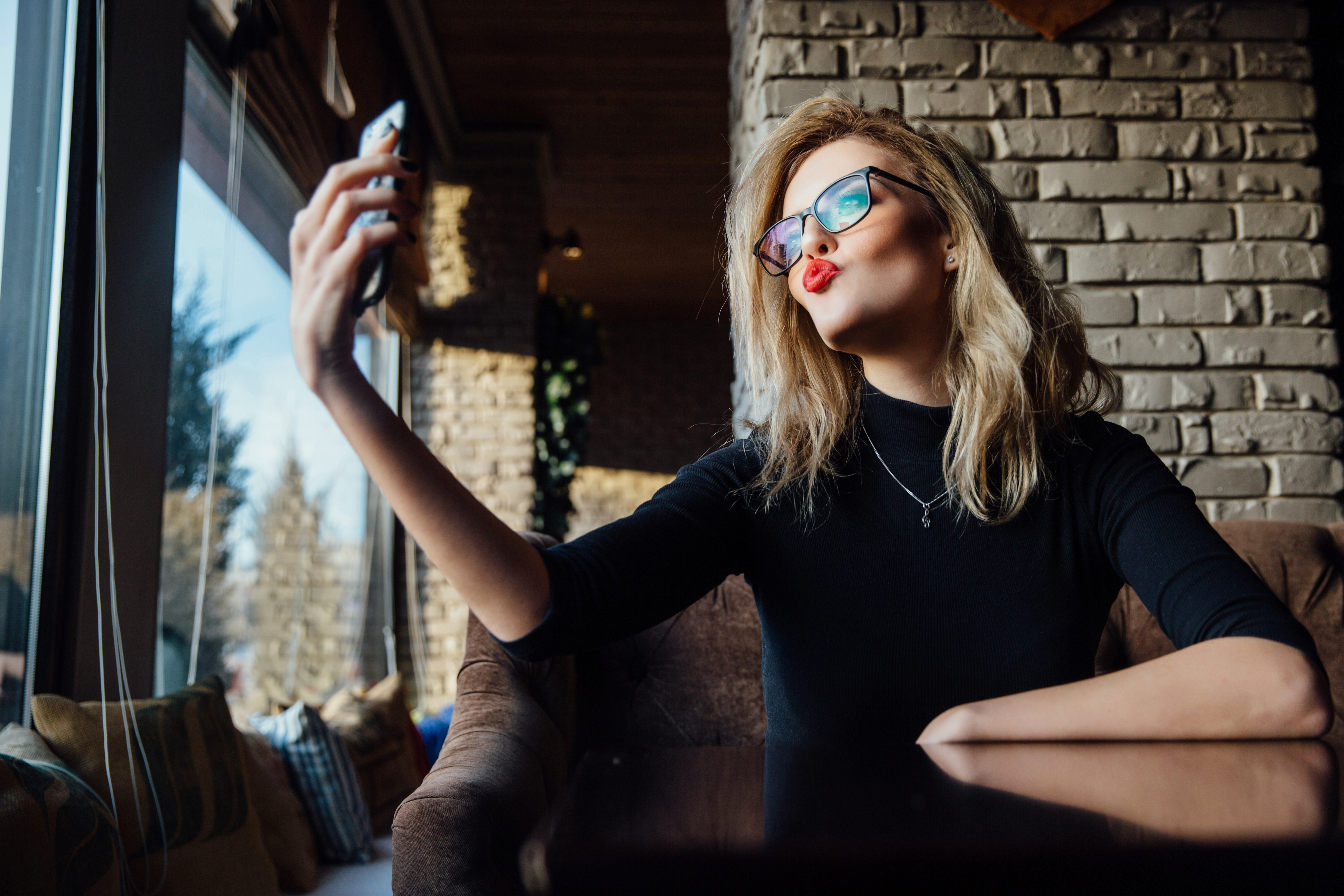 Young beautiful blonde woman taking a selfie in coffee shop. Hipster, red lips, glasses. Against the background of a brick stylish design wall.