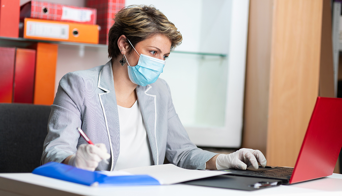 Mid adult woman using laptop and writing on the paper at work, wearing protective face mask and gloves against viruses