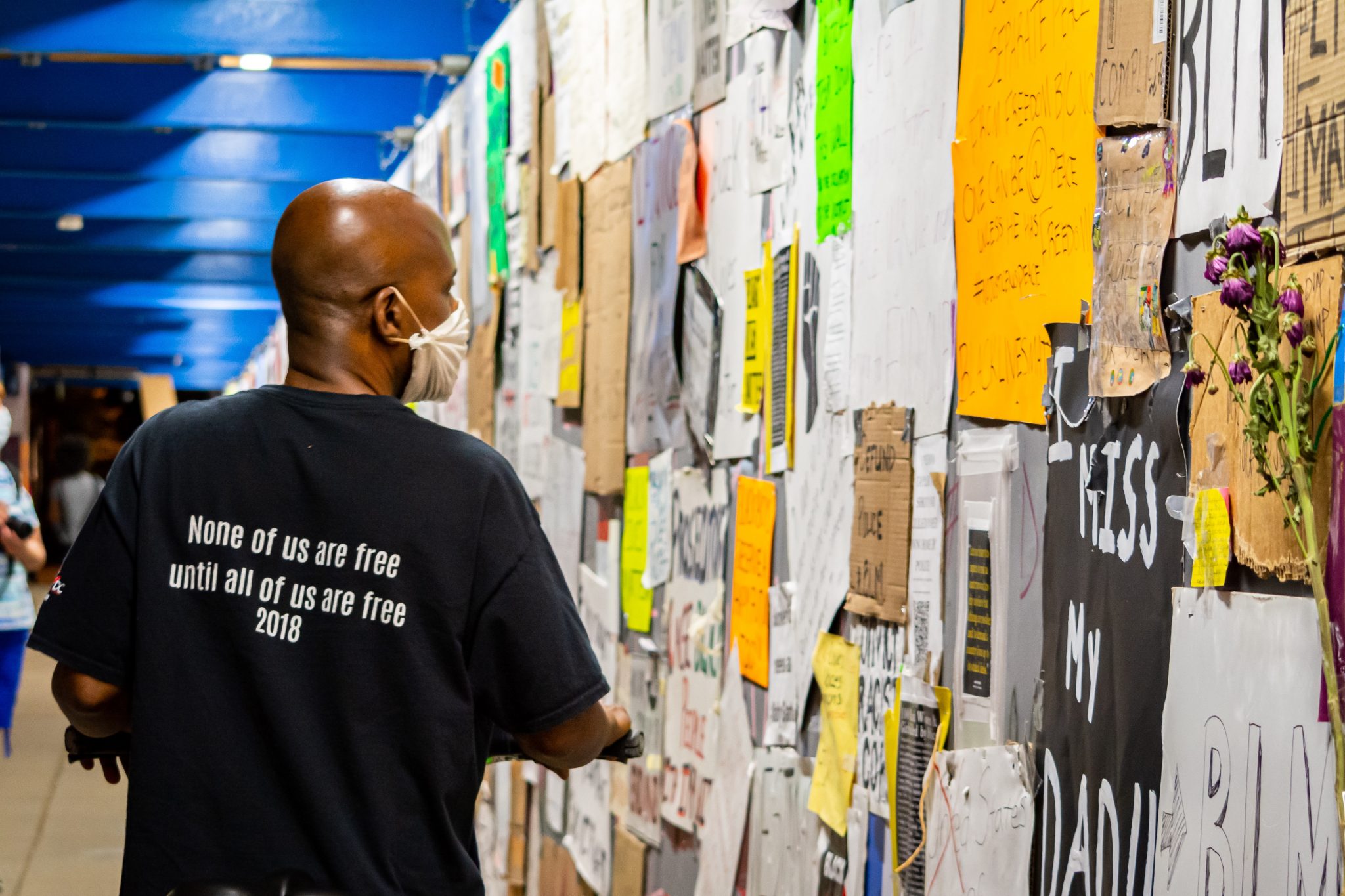 Photo of many in t-shirt that reads, &quot;None of us are free until all of us are free, 2018,&quot; looking at a bulletin board.