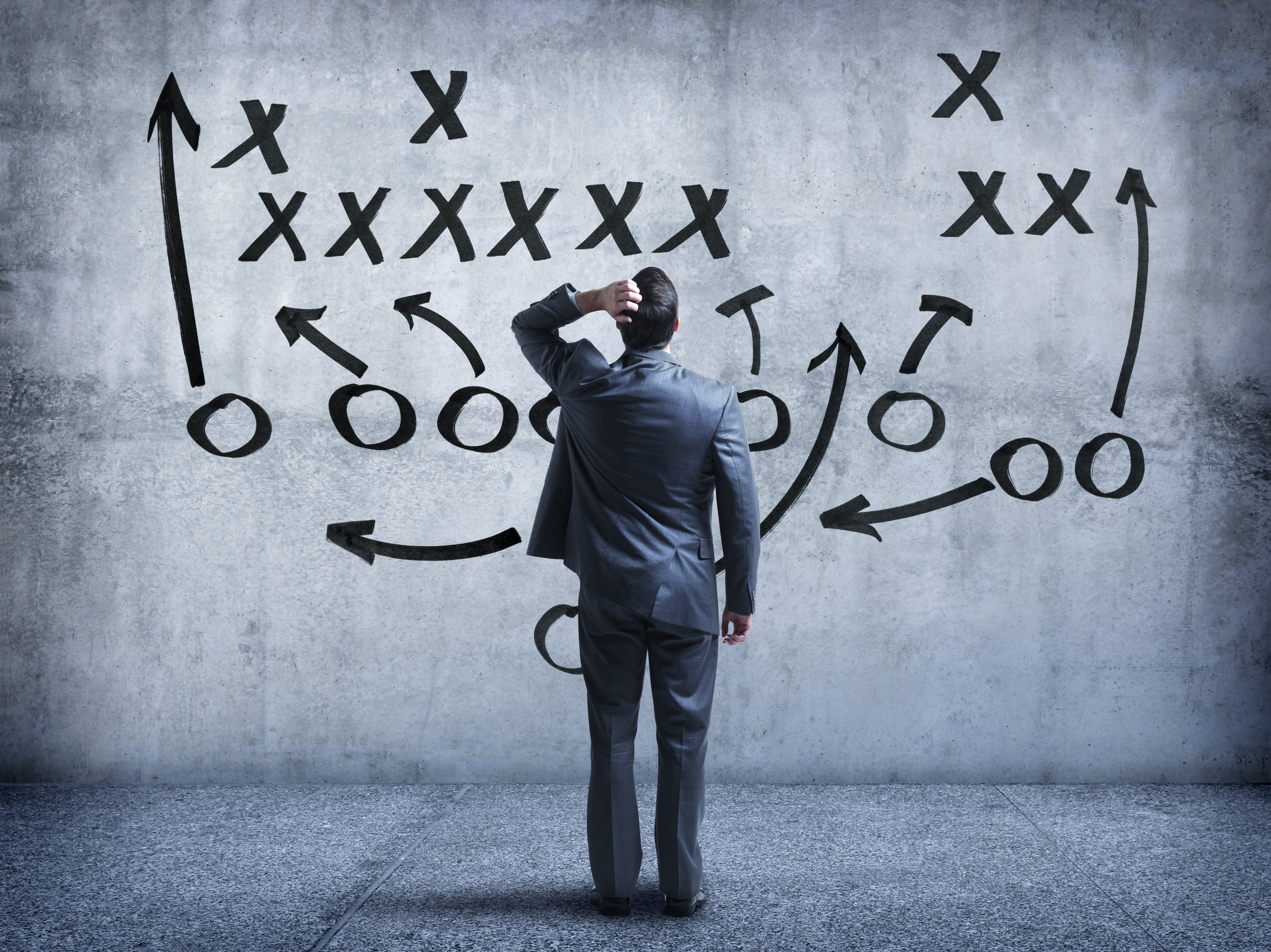 A rear view of a businessman looking up at an American football diagram written on a large concrete wall.  He is standing with his hand on his head as he tries to memorize all the different facets of the strategy. A blue color scheme dominates the image.